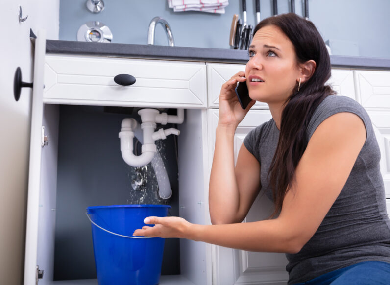 Worried Woman Calling Plumber While Collecting Water Leaking From Sink