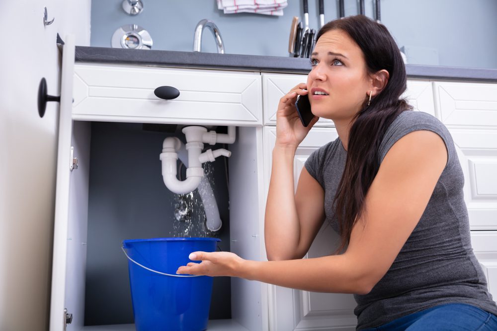 Worried Woman Calling Plumber While Collecting Water Leaking From Sink
